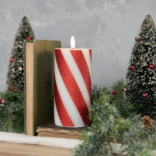 A 6-inch red 3D flame candy cane pillar candle with white stripes, placed on a table with holiday decorations around it, including a book, pine branches, and Christmas trees in the background.