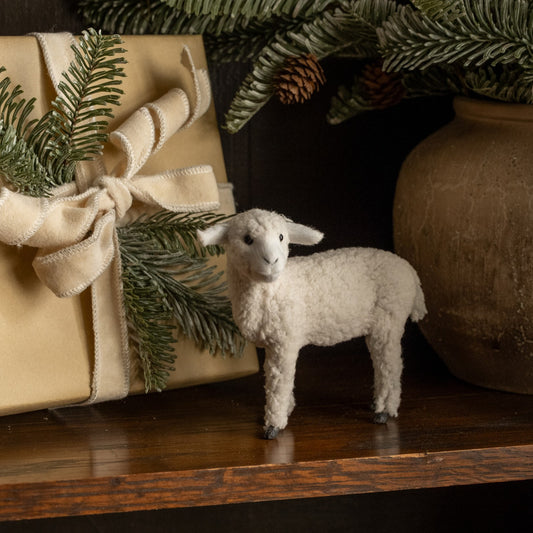 Small white lamb figurine next to a wrapped gift with greenery on a wooden surface.