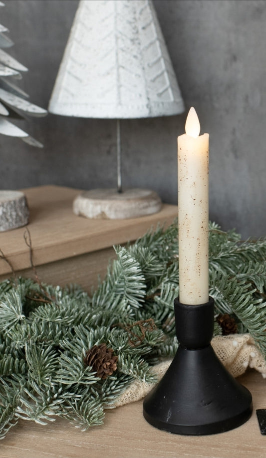 Candle in a black holder with a Christmas wreath and tree in the background