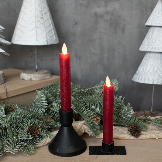 Two red candles in black holders with Christmas decorations on a wooden surface.