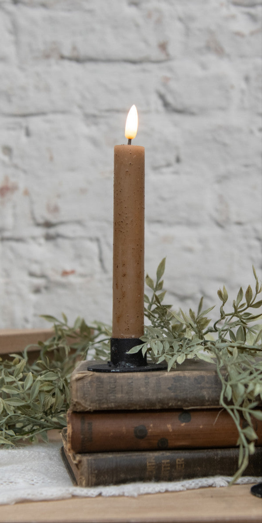 A beige taper candle with a 3D flame effect is lit and placed on a stack of old books, with a brick wall in the background and some greenery to the side.