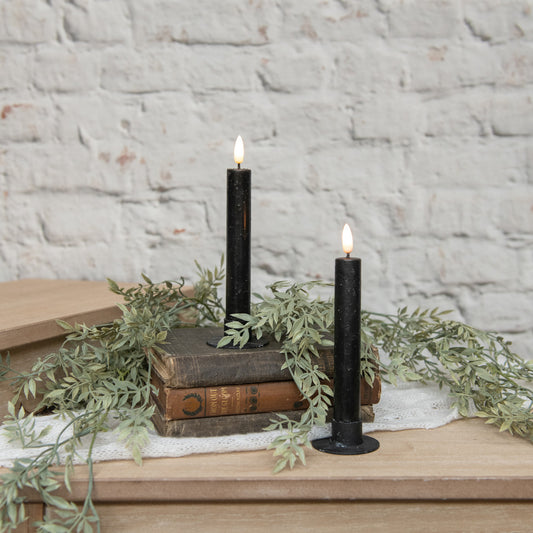 Two black candles on wooden books with greenery against a stone wall.