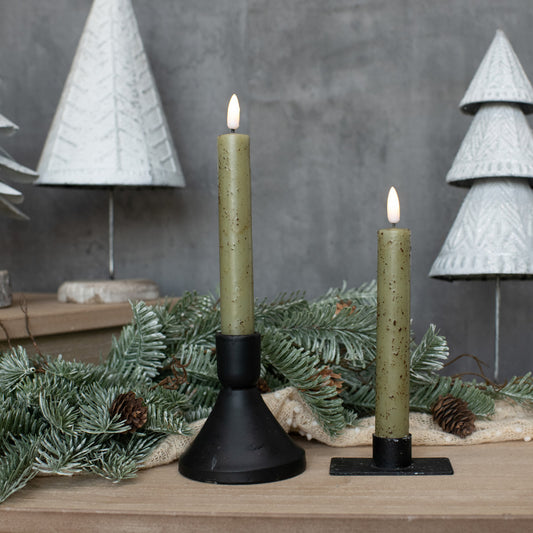 Two lit candles in black holders with Christmas decorations on a wooden surface.