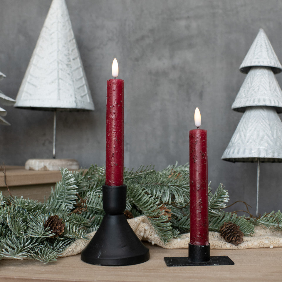 Two red candles in black holders with Christmas decorations on a wooden surface.