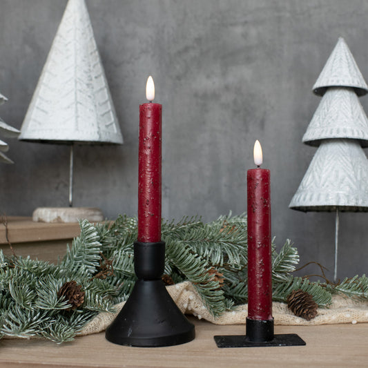 Two red candles in black holders with Christmas decorations on a wooden surface.