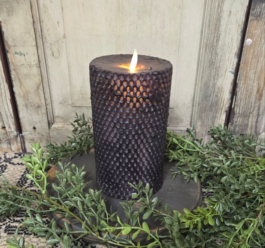 A black beeswax honeycomb pillar candle with a realistic moving flame, placed on a round metal holder, surrounded by green foliage.