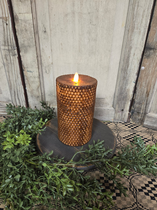 A battery-operated beeswax honeycomb pillar candle in brown color, displaying a realistic moving flame, placed on a surface with green foliage around it.
