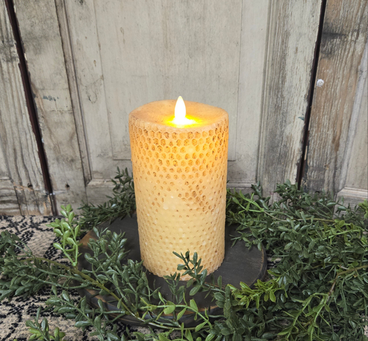 A 7-inch beeswax honeycomb pillar candle in burnt ivory color with a lit wick and a realistic moving flame, placed on a dark holder, surrounded by greenery.