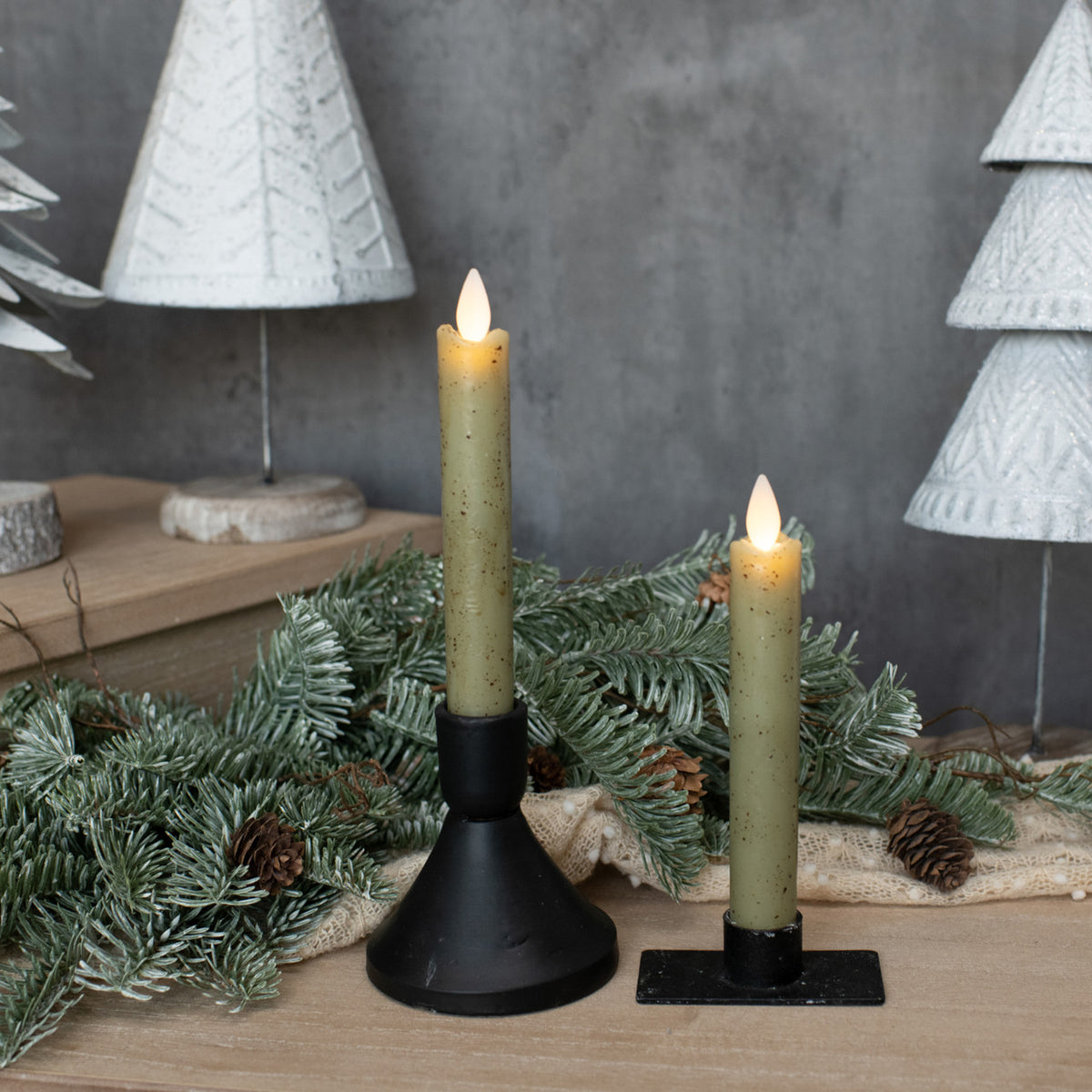 Two lit candles on black stands with Christmas decorations in the background.