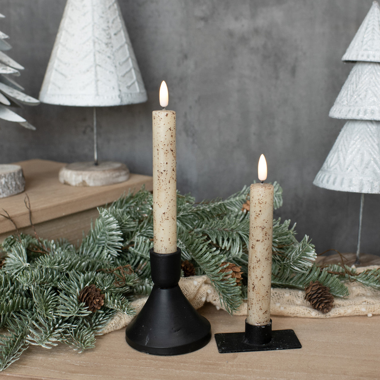 Two lit candles on black stands with decorative greenery and white textured trees in the background.