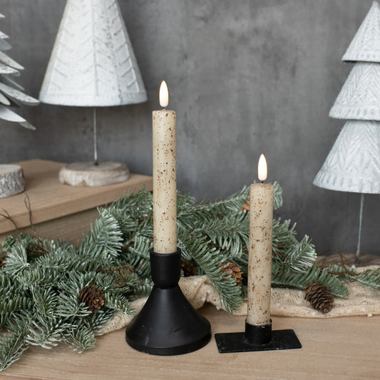 Two lit candles on black stands with decorative greenery and white textured trees in the background.
