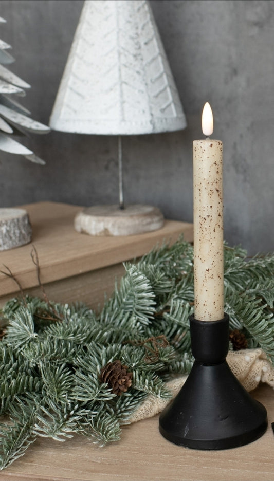 Candle in a black holder with a Christmas tree and decorative items in the background