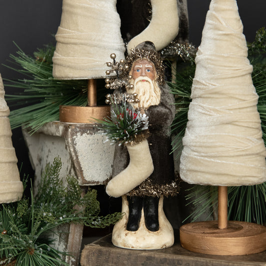 Decorative figure of a bearded man holding a stocking with Christmas trees and greenery in the background.