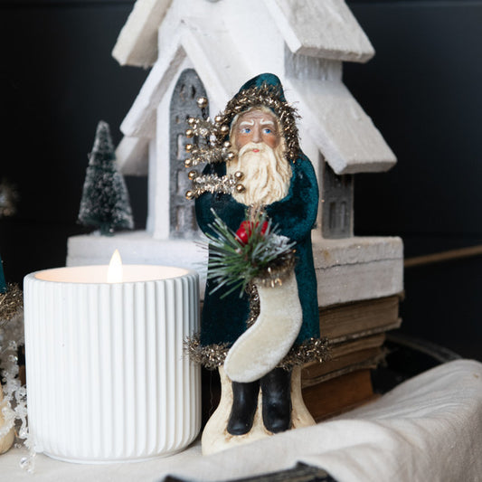 A decorative figurine of a bearded man in a blue velvet suit holding a holly wreath, standing next to a white candle on a mantel.