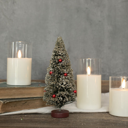 A small artificial green bottlebrush tree with red berries, placed between two candles on a wooden surface, with a gray wall in the background.