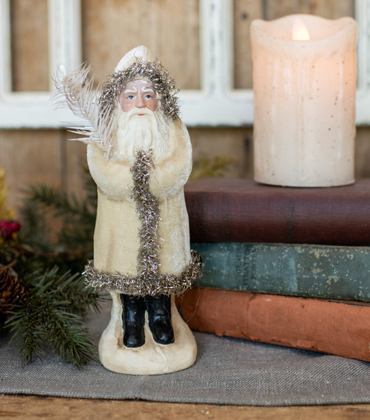 A Santa Claus figurine in a cream-colored coat with silver tinsel trim, a white beard, and black boots, standing on a base. It is styled on a tabletop next to pine branches and a stack of books with a lit candle in the background.