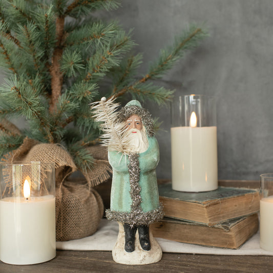 An 8-inch soft blue velvet Belsnickle figure, resembling a Santa Claus-like character, standing on a small base, with a backdrop of a pine tree and lit candles.