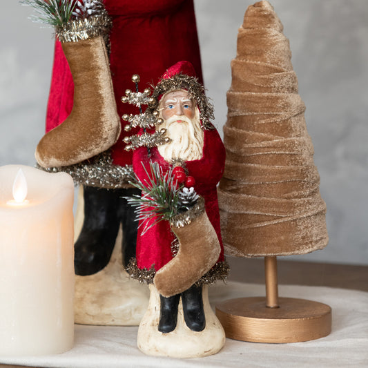 Wine colored Santa with beard holding a tan socking with velvet tree beside it.