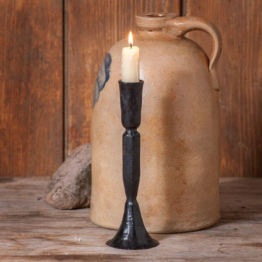 A black cast iron taper candle holder with a lit candle, placed on a wooden surface next to a beige ceramic jug and some rocks.