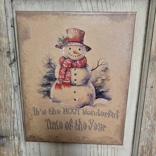 A canvas print featuring a snowman with a red hat and a green scarf, sitting on a wooden surface with the text 'It's the most wonderful time of the year'.