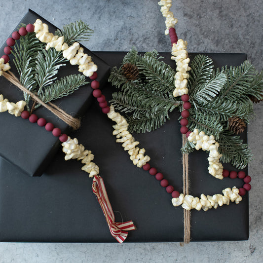 A decorative garland made with artificial cranberries and popcorn, tied with twine and adorned with ribbons, presented in front of a black gift box.