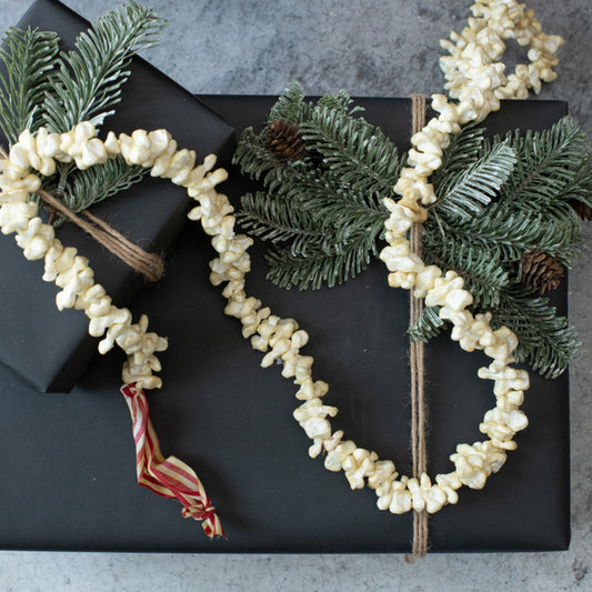 A 9-foot long popcorn garland made from artificial popcorn, displayed alongside green faux pine branches and red ribbon accents.