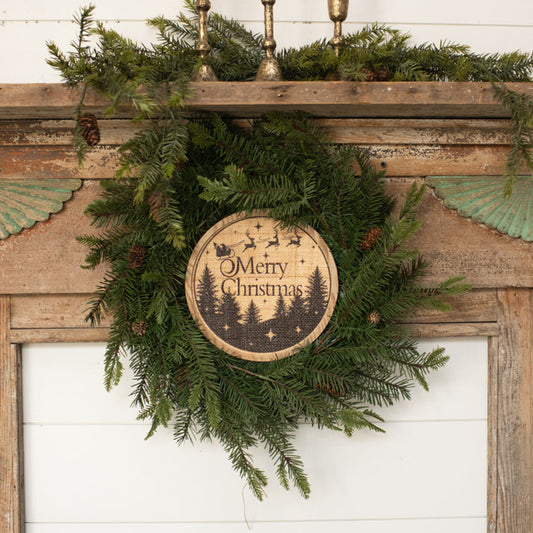 A round 'Merry Christmas' sign with a festive design, hanging on a wooden surface, featuring greenery and a circular brown background with the text 'Merry Christmas' in the center.