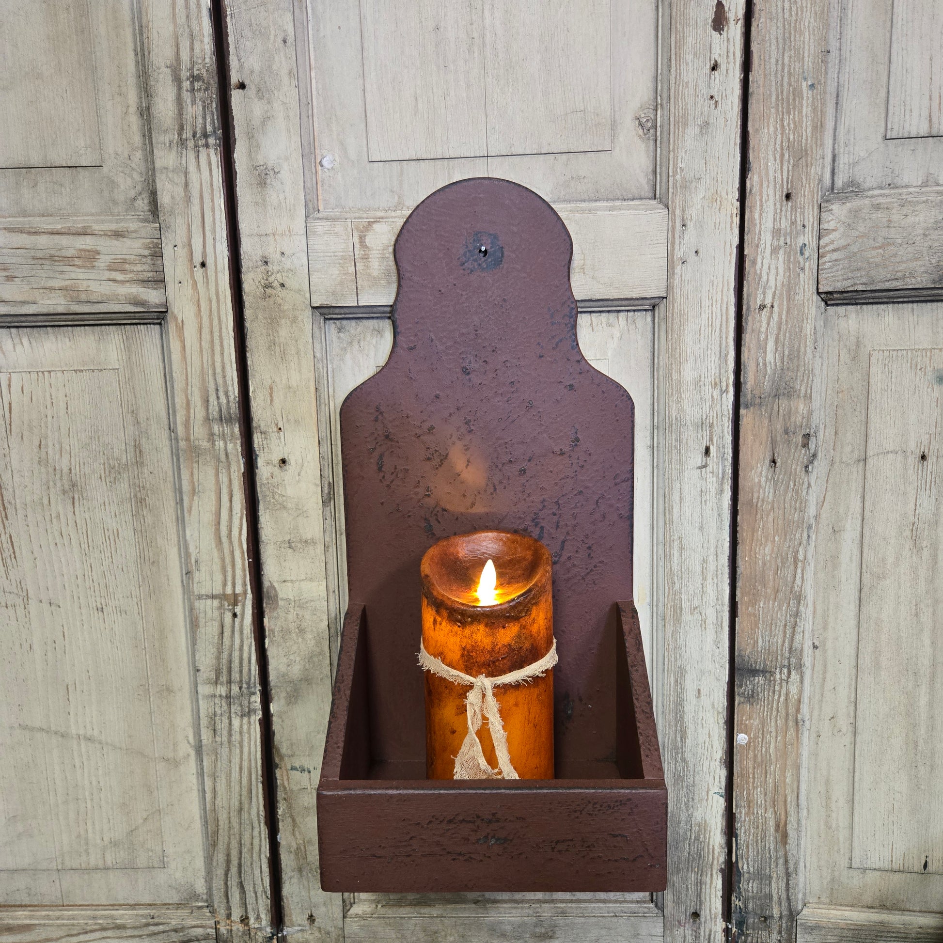 A burgundy wooden sconce candle holder mounted on a wall, with a candle inside it, placed against a rustic wooden background.