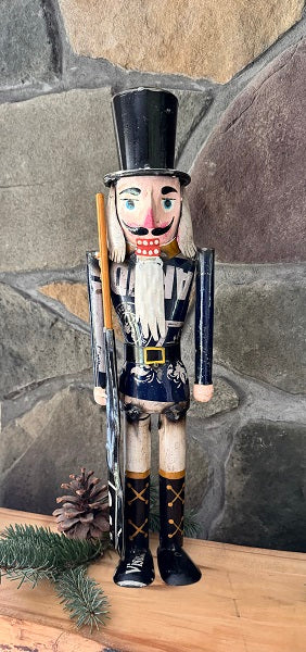 A decorative metal nutcracker in a vintage style, predominantly blue in color, with a black hat and white facial features, standing against a stone background with pine cones and greenery.