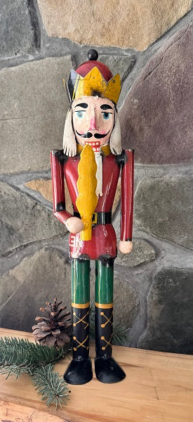 A decorative metal nutcracker with a vintage design, featuring a red jacket, green pants, black boots, and a yellow scarf, positioned against a stone background with pine cones and a Christmas tree branch.