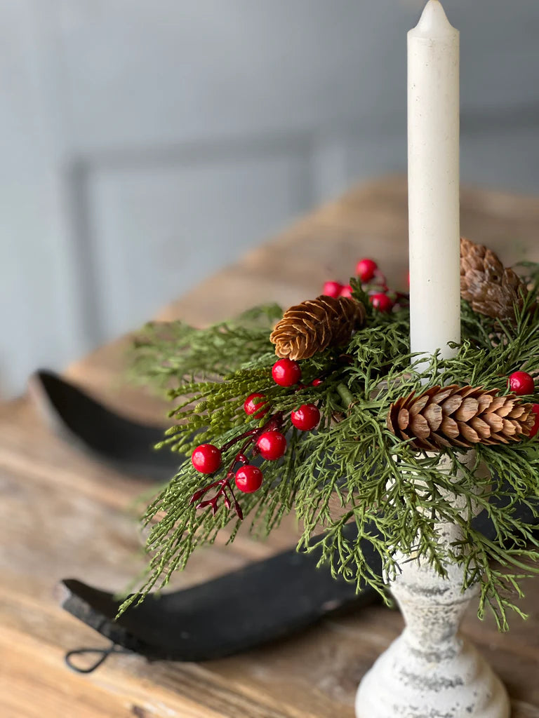 Decorative candle holder with greenery, red berries, and pine cones on a wooden surface.