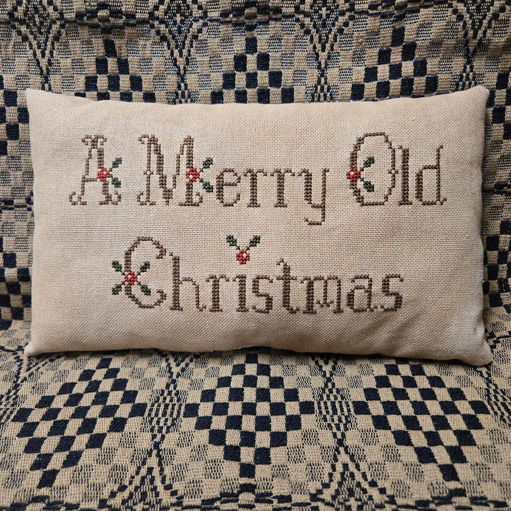 A rectangular beige linen pillow with 'A Merry Old Christmas' stitched in brown thread with small red and green holly accents, resting on a black and cream checkered patterned couch.