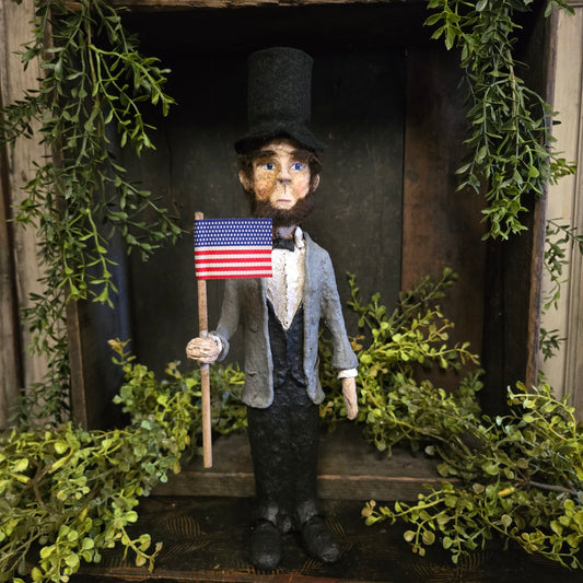 Small figurine of a person holding an American flag, surrounded by greenery.