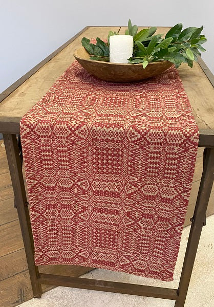 A red and beige geometric patterned table runner displayed on a wooden table with a decorative bowl and greenery on top.