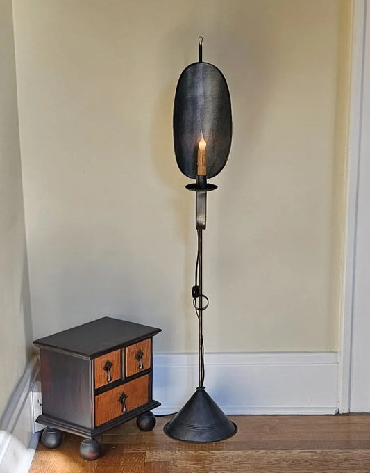 A mini floor lamp with a rustic metal finish mounted on a wooden floor next to a small wooden cabinet. The lamp has a warm light illuminated at the top.