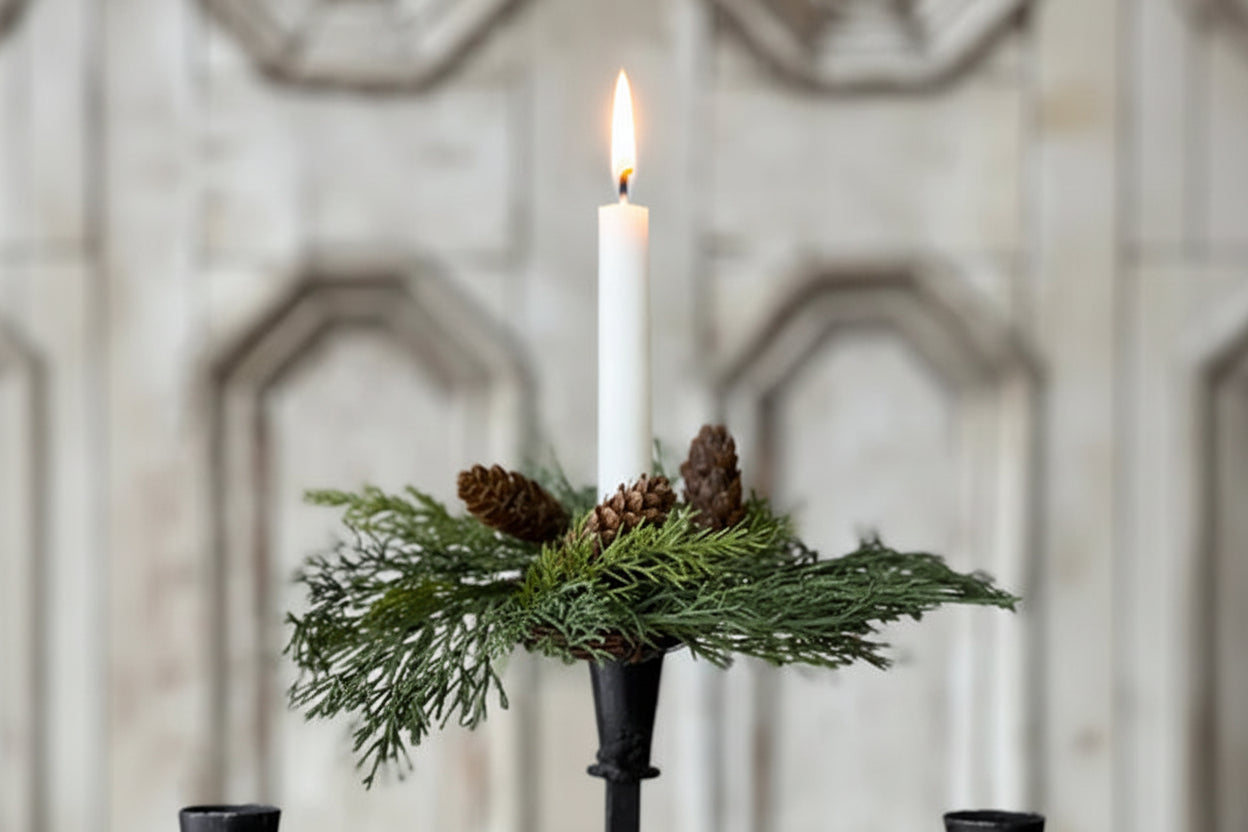 Decorative candle holder with greenery and pinecones against a decorative wall.
