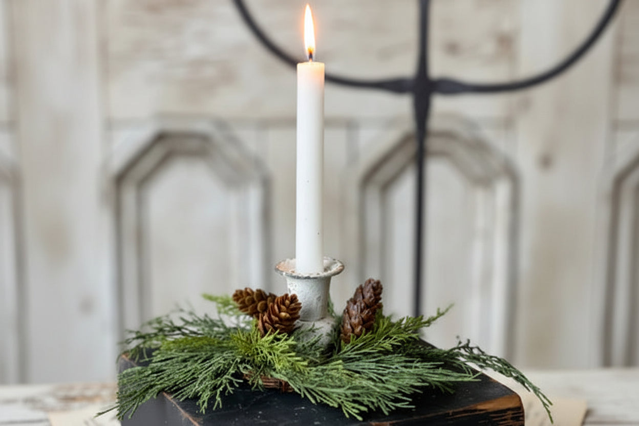 A rustic holiday centerpiece featuring a white taper candle in a distressed white metal holder, nestled in green pine branches and small pinecones atop a dark wooden block, with a blurred vintage-style background.