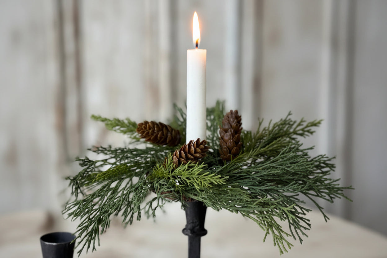 A white taper candle in a black metal holder, surrounded by green evergreen branches and three brown pine cones, set against a softly blurred light gray background.