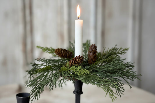 A white taper candle in a black metal holder, surrounded by green evergreen branches and three brown pine cones, set against a softly blurred light gray background.