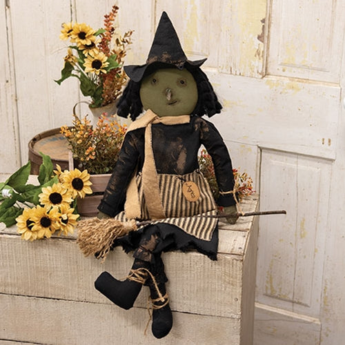 Decorative witch figurine with a broom in a rustic setting with flowers and a wooden surface.