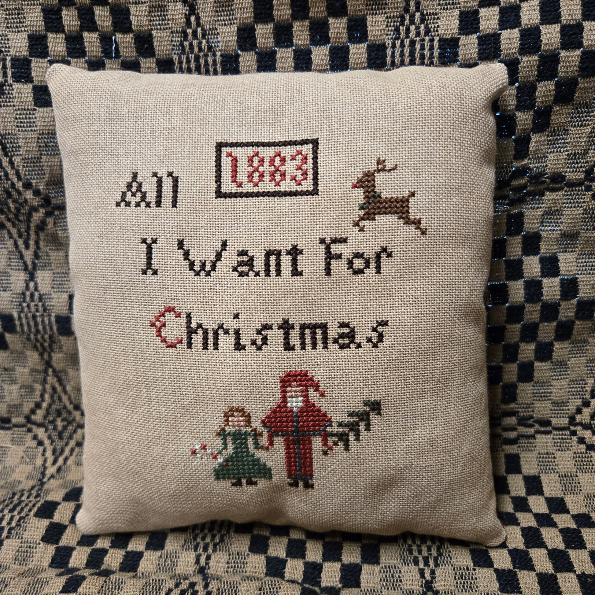 A square beige linen cross-stitch pillow with embroidered Christmas text and figures, placed on a black-and-white checkered blanket. The embroidery reads 'All I Want For Christmas' with '1883' in a box, plus a reindeer, Santa, and children.