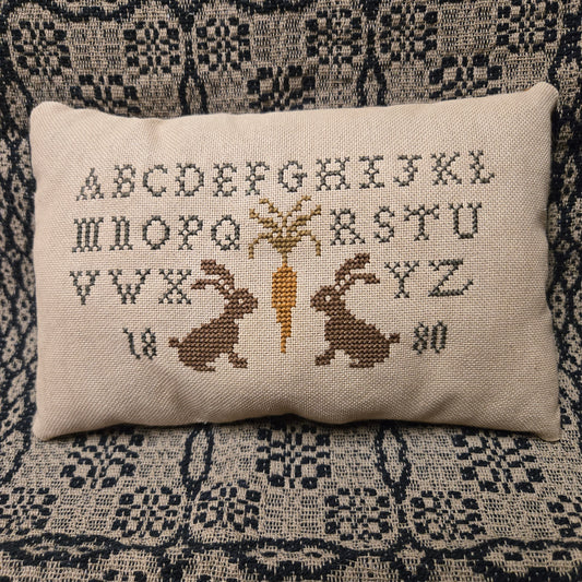 Decorative pillow with cross-stitched designs of a bunnies and carrots on a patterned fabric background.