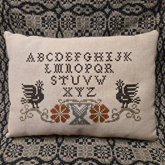 Decorative pillow with cross-stitched design featuring flowers and letters on a textured fabric background.