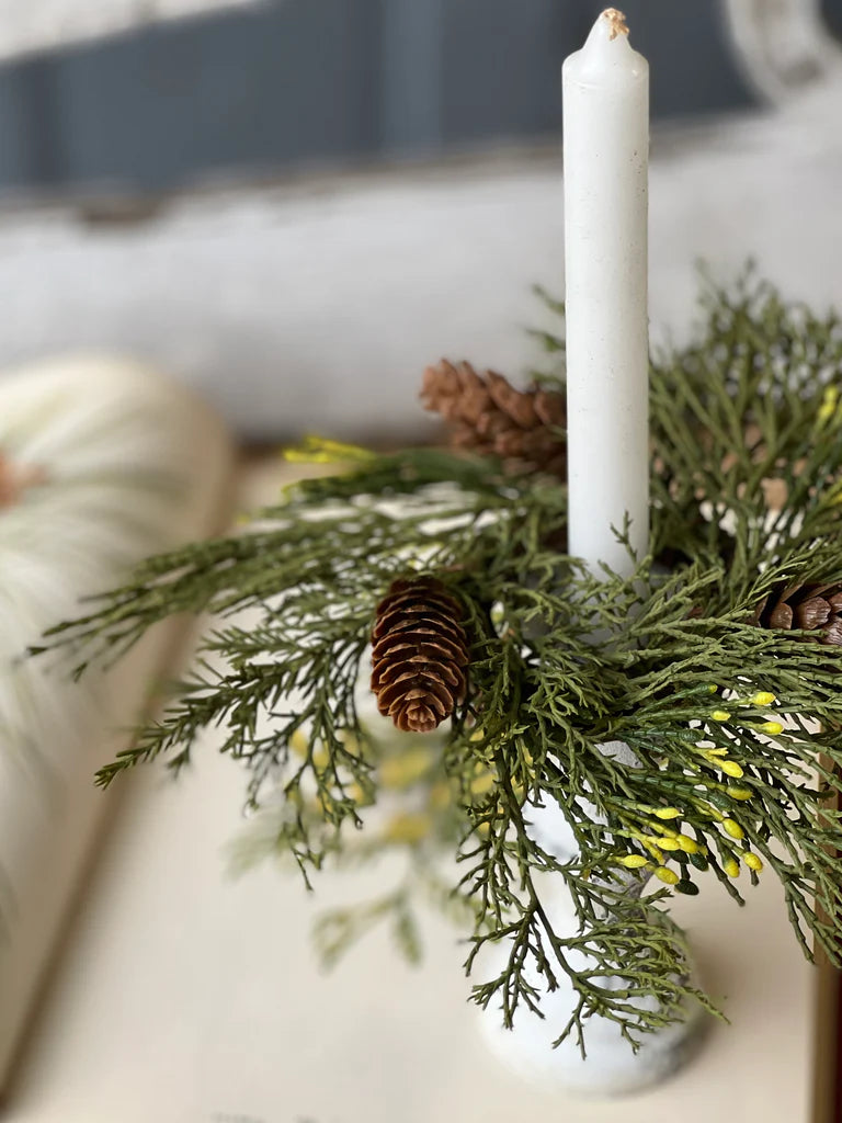 White candle in a holder with greenery and pine cones on a neutral background
