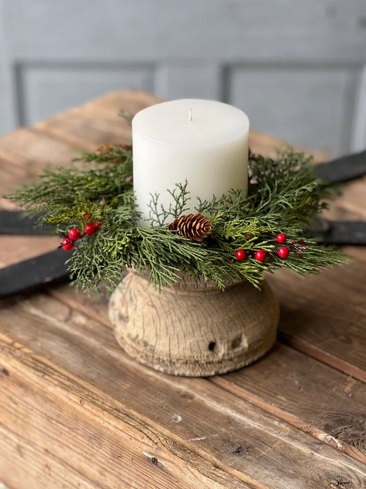 A candle ring decorated with green cedar, red berries, and pine cones, displayed on a wooden surface.