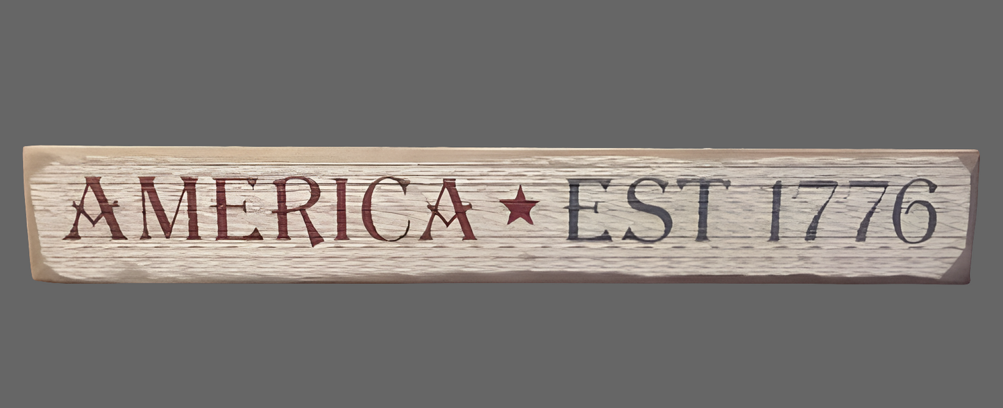 A wooden sign with the text 'AMERICA EST. 1776' in white and red, with a star symbol, on a wooden surface.