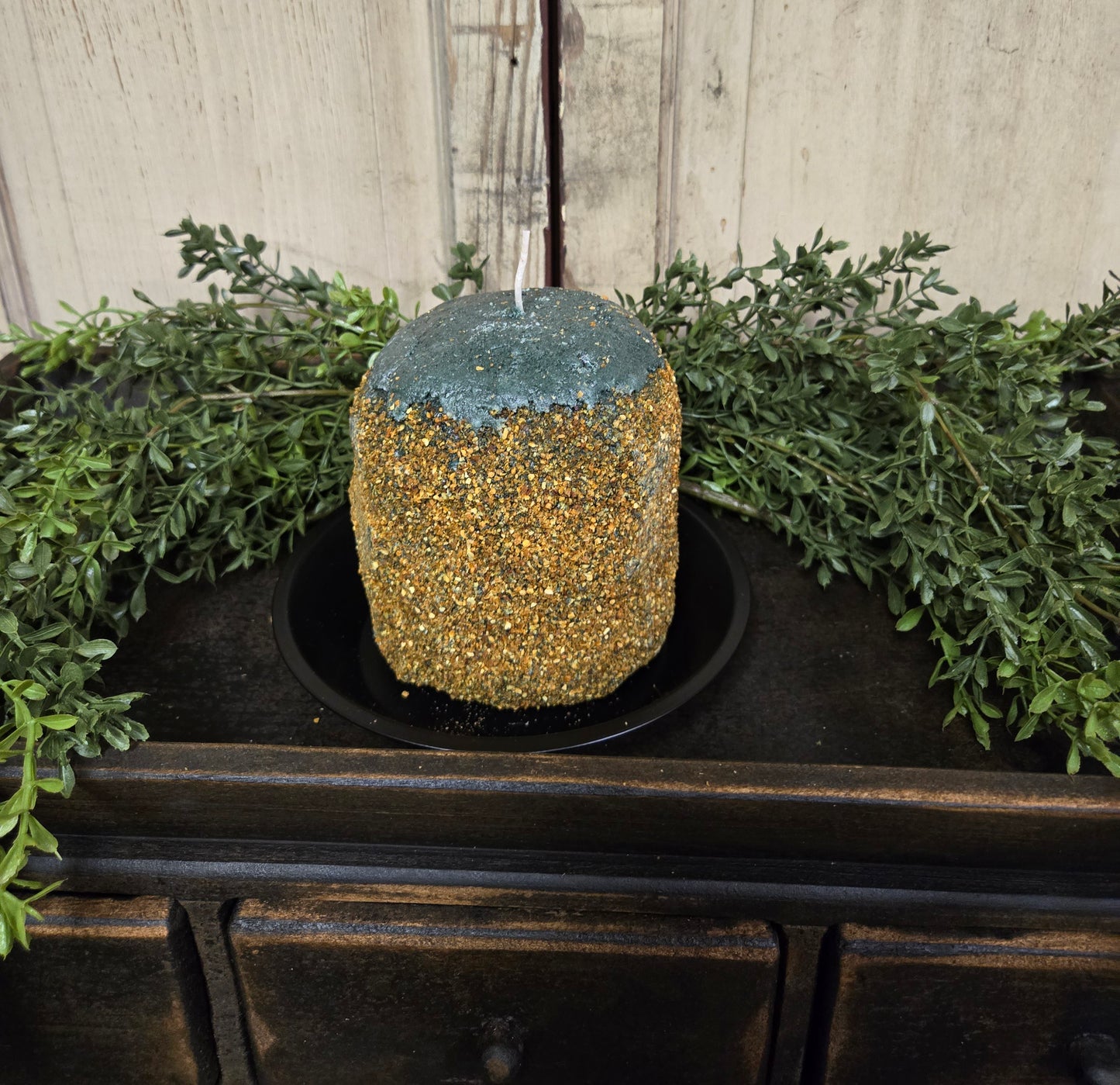 A green and brown hearth candle with a primitive design, placed on a black dish, surrounded by greenery and set against a wooden background.