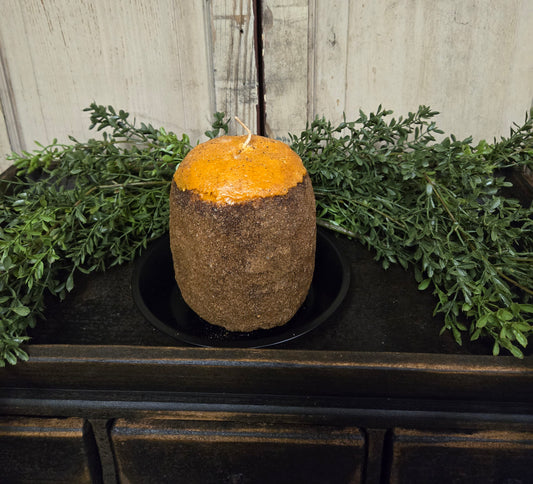 A cylindrical gingerbread Hearth candle displayed on a hearth, surrounded by greenery. The candle has a brown exterior with a golden yellow top and is partially consumed, showing a visible wick.