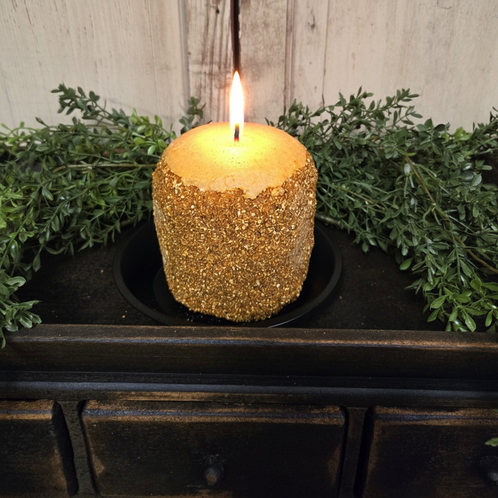 A lemon custard-scented hearth candle placed on a black dish, surrounded by greenery and set against a rustic wooden background.