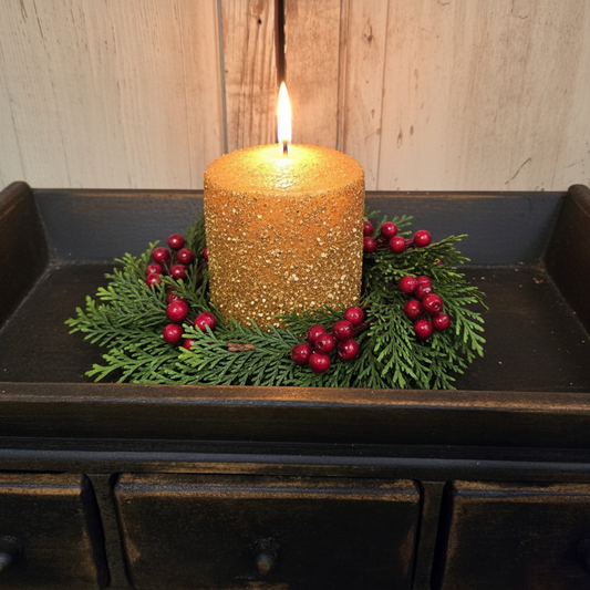 Gold glitter candle with greenery and red berries on a dark surface.